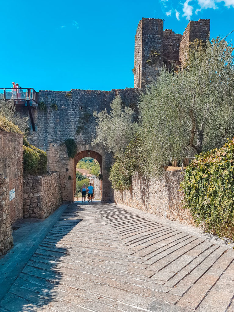 Una porta d'ingresso al castello di Monteriggioni costeggiata da muriccioli bassi con ulivi. Sopra alla porta si trova il camminamento delle mura e sulla destra un torrione difensivo.