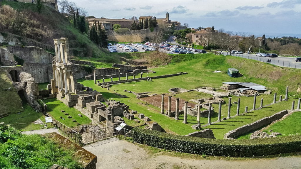 L'antico anfiteatro romano di volterra visto al completo con in primo piano le colonne rimaste in piedi e parte del frontescena. Tutto inserito in un prato verde.