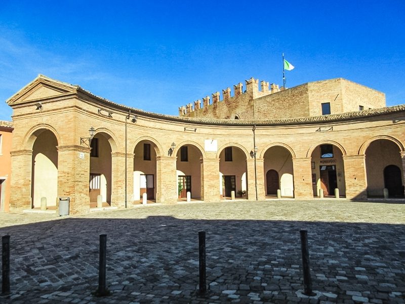 La piazza principale di Mondaino con la sua forma ovale e arcata come se stesse abbracciando il visitatore. Sullo sfondo in alto si staglia un antica fortificazione medievale con la merlatura.