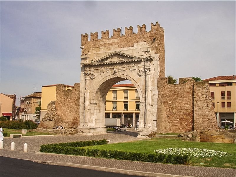 L'arco di Augusto a Rimini con la sua merlatura medievale e parte delle fortificazioni ancora in piedi. Si tratta di un antica porta di ingresso al centro storico e posta davanti ad un praticello verde.