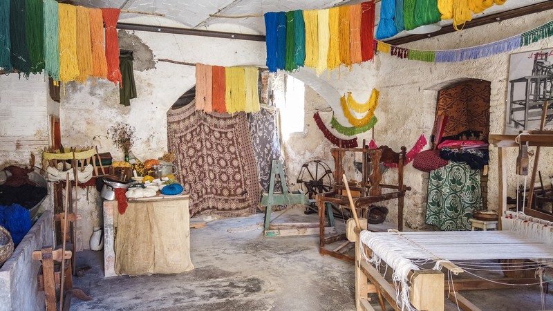 Gli antichi strumenti e macchinari di tessitura di Rasiglia Umbria. In primo piano una tessitrice e un tavolo con altri strumenti. Appesi sul soffitto i tessuti di varie colorazioni.