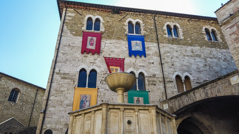Un primo piano sul Palazzo dei Consoli di Bevagna con le sue finestre biforate e alcuni stendardi colorati appesi. Frontalmente si trova la fontana medievale