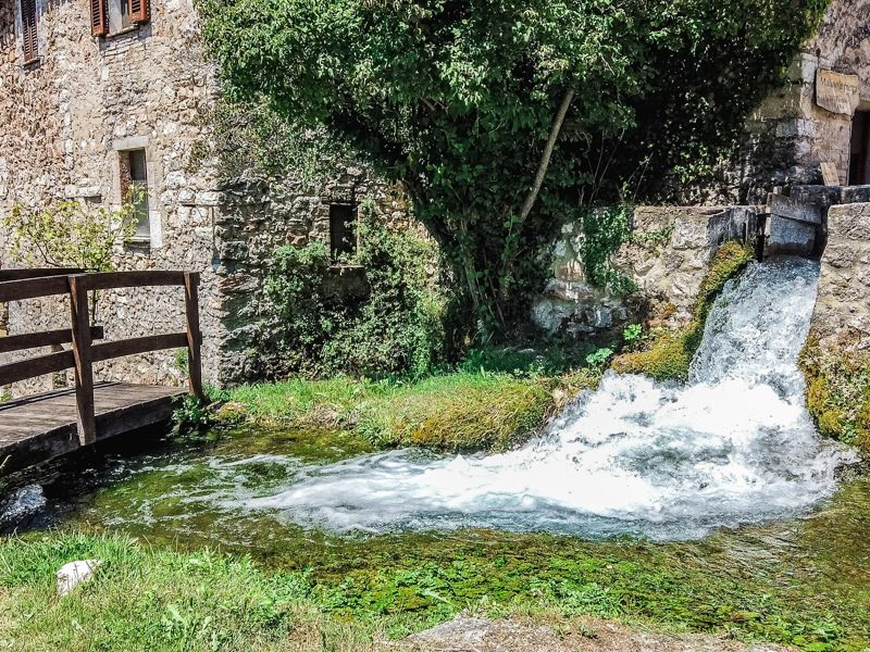 Una piccola cascata d'acqua che sfocia in un ruscello all'interno del borgo di Rasiglia. Sulla sinistra un ponticello in legno che fa attraversare il visitatore alla sponda opposta.