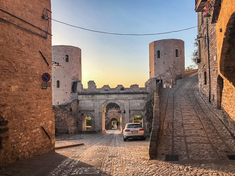 Una delle porte d'ingresso al borgo di Spello in provincia di perugia. La porta è sotto un grandissimo arco con due alti torrioni medievali ai lati. In primo piano invece due stradine ciottolate medievali.