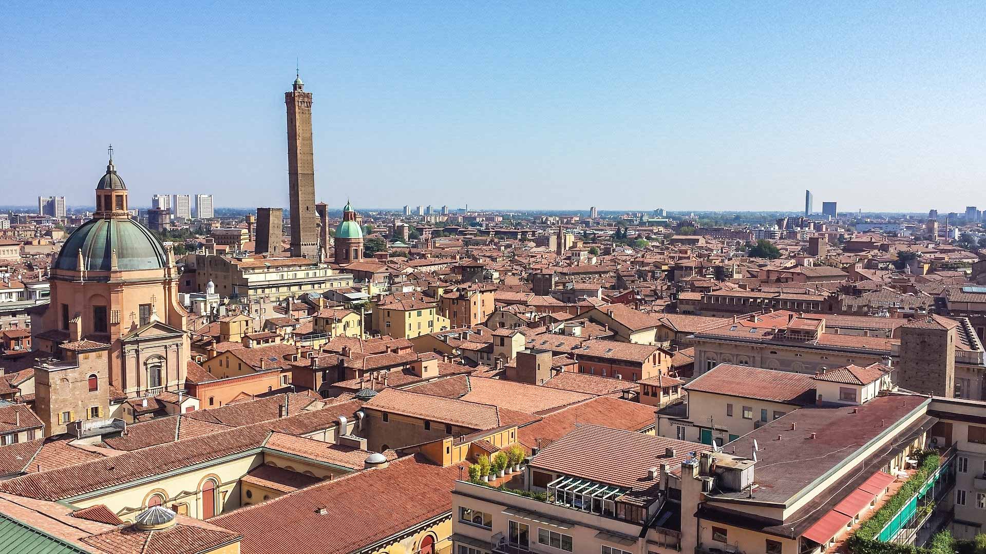 Una vista spettacolare dall'alto su tutta la città di Bologna. Tra i tanti edifici spiccano la cupola di una chiesa e soprattutto l'altissima torre degli asinelli. Sullo sfondo gli edifici moderni della città.