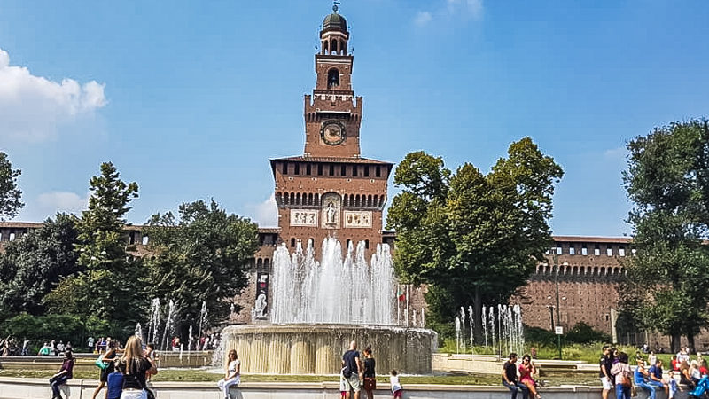 Il Castello Sforzesco è l'attrazione più importante da vedere a Milano in un giorno. Sullo sfondo l'altissima torre con orologio e alcuni rilievi in marmo mentre in primo piano la fontana con i giochi d'acqua.