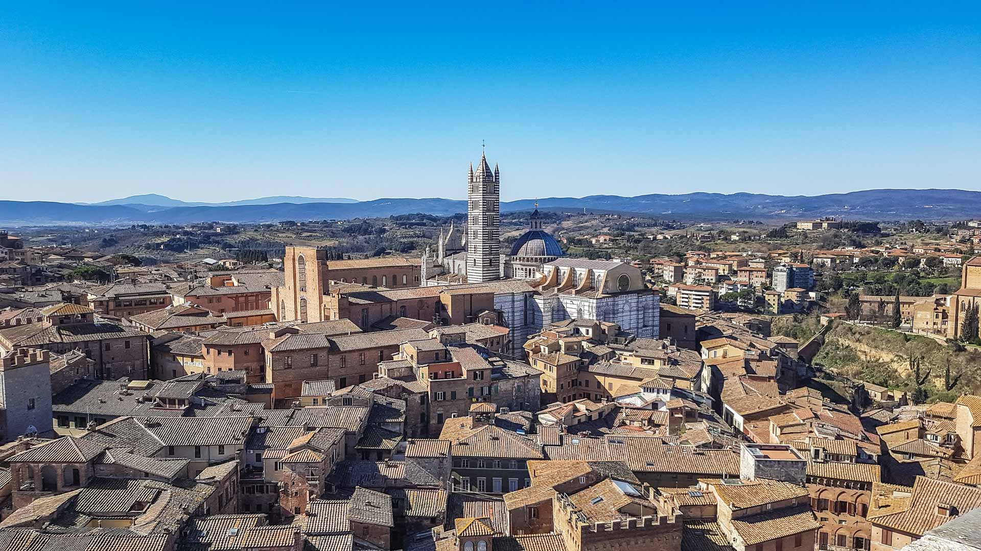 Una meravigliosa vista completa su tutta Siena con al centro della foto la bellissima cattedrale. Sullo sfondo colline e montagne. La città è sicuramente una delle mete da vedere in un giorno di vacanza!