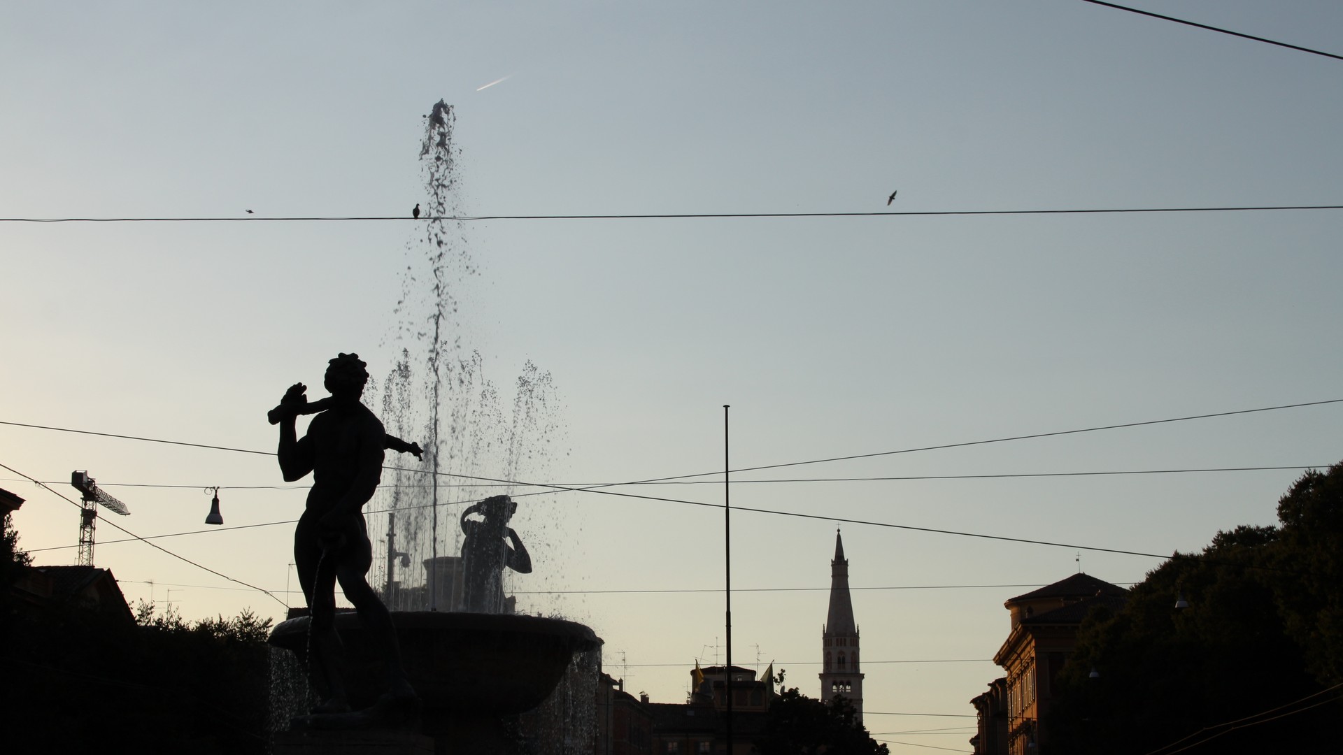 Un bellissimo primo piano su una delle fontane più belle di tutta Modena con le statue in risalto davanti e dietro lo spruzzo d'acqua. Sullo sfondo si vede il campanile a punta di una chiesa.