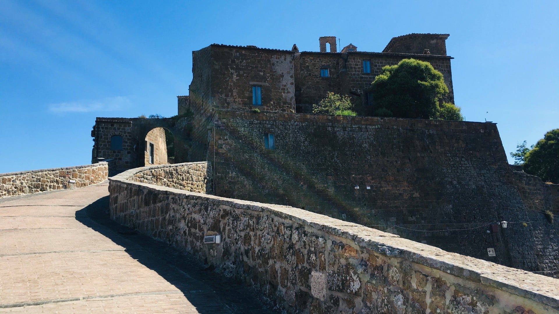 Una passeggiata che accompagna il turista all'entrata del borgo fantasma di Celleno. Si vede sullo sfondo l'arco dell'entrata al borgo e accanto spiccano le case in mattoni.