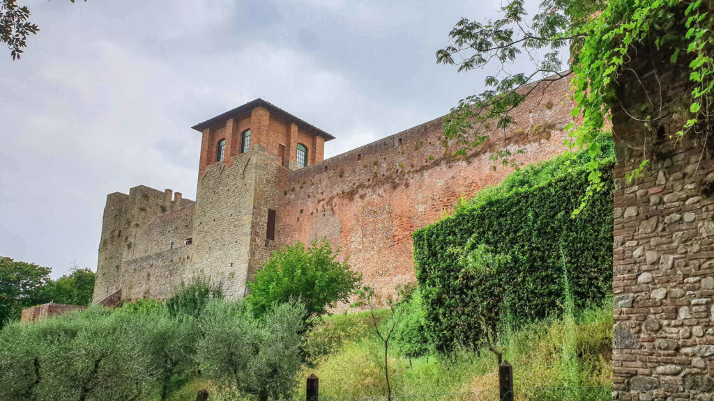 Le alte mura in pietra della Fortezza del Cerruglio di Montecarlo di Lucca. In primo piano si vedono i due torrioni quadrati con sotto alcuni ulivi e cespugli verdi.