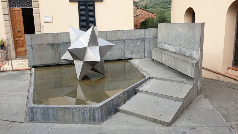 Un particolare della piazza dei guidi nel paese vinci. Si nota una forma geometrica in ferro nel mezzo di una piccola vasca d'acqua con accanto due grandi scalini in pietra.