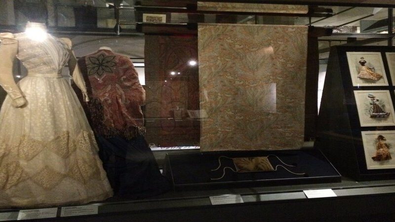 una vetrina del museo del tessuto di prato che espone due abiti antichi molto sfarzosi ed un tessuto. Sulla destra si intravedono anche alcuni disegni di altri abiti storici.