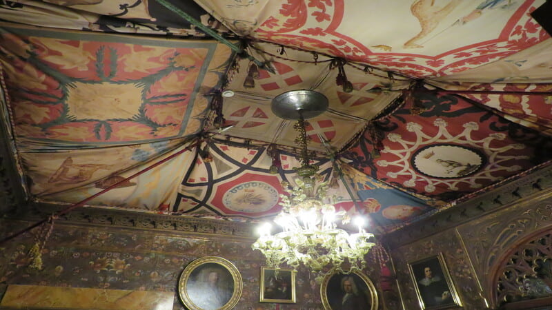 un primo piano sulle bandiere del palio di siena comprate da stibbert a siena. Al centro del soffitto un lampadario antico che illumina le bandiere.