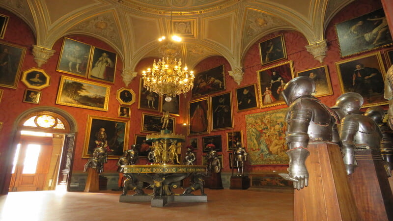 la sala della malachite del museo stibbert di firenze con i suoi tantissimi quadri appesi al muro e alcune armature medievali agli angoli. Al centro della stanza un grande tavolo e un lampadario antico.