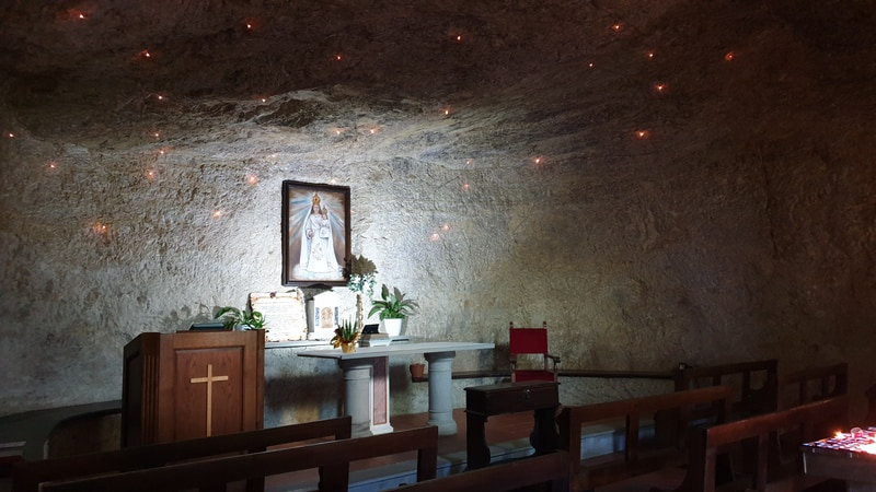 l'interno della sacra grotta dell'eremo di calomini con in fondo al centro la sacra immagine della vergine e alcune panchine. il soffitto roccioso è decorato con lucine che sembrano stelle luminose