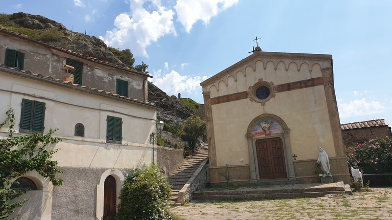 in primo piano la facciata spoglia della chiesa di sant'andrea di montemassi, sulla destra una salita che porta verso altre case del centro storico di montemassi mentre in alto si intravede parte della rocca medievale