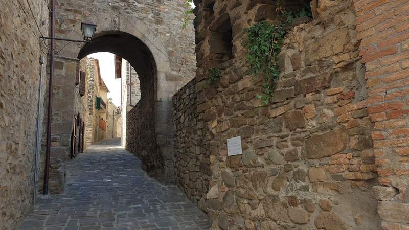 un antica strada in mattoni contornata da mura antiche in pietra sul cui sfondo si trova un'alta porta d'ingresso al centro storico di montenero d'orcia