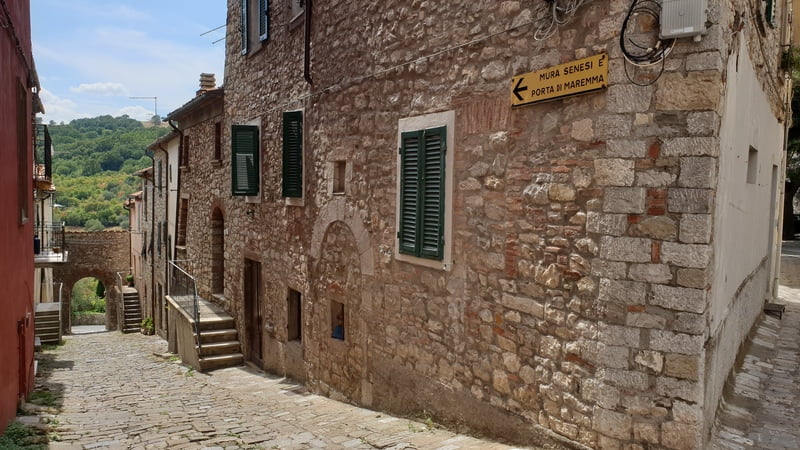 una lunga strada medievale contornata da edifici in pietra dove in fondo si vede un antica porta di ingresso al centro storico di roccalbegna a forma di arco