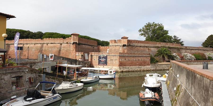 una vista in primo piano sulle imponenti mura marroni di mattoni della fortezza nuova di livorno con in basso il canale di acqua e alcune barche ormeggiate nei vari pontili in legno
